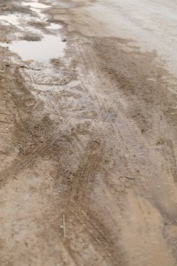 Çamurlu bir patikada yağmurdan sonra lastik izleri ve su birikintileri görülüyor. Zemin ıslak ve düzensiz, son hava değişikliklerini yansıtıyor. Bu sahne kırsal bölgelerde yaygındır..