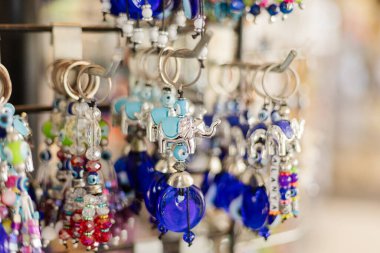 Karmaşık fil tasarımlı anahtarlıklar ve renkli boncuklar bir marketin vitrininde asılı duruyor. Parlak maddeler, insanlar etrafa göz attıkça ışığı yansıtıyor.