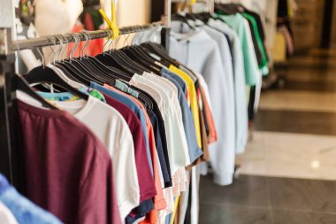 Perakende mağazasındaki askılarda farklı renk ve tarzlarda gömlekler düzenlenir. Sahne hareketli, müşteriler etrafta dolanıp bakınıyor..