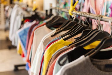 Bir perakende mağazasının içinde çeşitli renklerde tişörtler asılıdır. Alışverişçiler mağazanın yoğun atmosferinin keyfini çıkarırken giysilere göz atıyorlar..