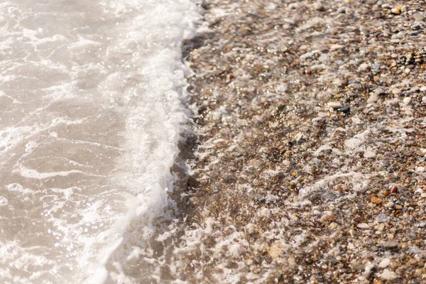 Dalgalar küçük taşlar ve çakıl taşlarıyla kaplı bir sahile çarpar. Su girip çıkıyor, kıyıda desenler oluşturuyor. Güneş ışığı ıslak taşlara yansıyor..