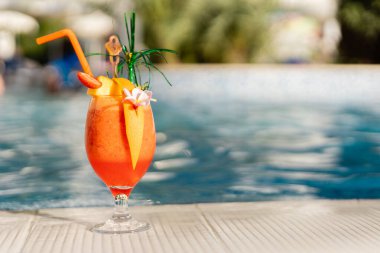 Parlak bir içecek havuzun yanındaki beyaz bir yüzeye konur. İçki meyve ve çiçeklerle süslenmiştir. Güneş ışığı suyu yansıtır ve rahatlatıcı bir atmosfer yaratır..