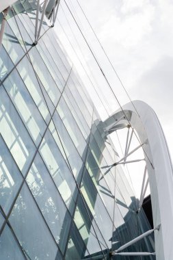 Şehirdeki inşaat, cam duvarları ve eşsiz bir yapısı olan modern bir bina gösteriyor. Mimarlar binayı dikkat çekmek için tasarladılar. Bulutlar gökyüzünü doldururken insanlar yakınlarda yürür.