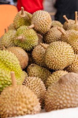 A large group of durian fruits is arranged in a market setting. Some fruits have a greenish hue while others are more yellow. People are seen in the background.