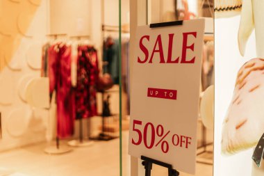 Bir tabela bir giyim mağazasında yüzde 50 indirim olduğunu gösteriyor. Renkli giysiler arka plandaki raflarda asılı dururken, alışveriş yapanlar yakınlarda geziniyor..