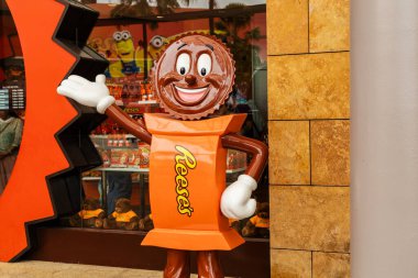 Singapur - 7 Ocak 2026: Çikolata ve fıstık ezmesi gibi giyinmiş bir karakter ziyaretçileri bir mağazanın önünde karşılıyor. Reese 'in logosu. Karakter renkli bir ekranın önünde duruyor..