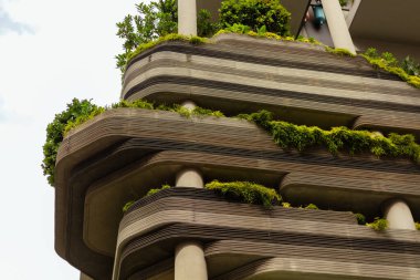 Benzersiz tasarım özellikleri olan bir binanın yakın görüntüsü. Kenarlarında yeşillikler ve bitkiler olan birkaç balkon var. Gökyüzü açık ve ayara parlak bir dokunuş katıyor..