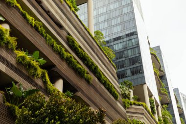 Modern bir yapı, bitkilerle kaplı birden fazla katmanı olan uzun bir yapıdır. Dekor, beton ve yeşillik karışımı içeriyor. Görüntü yoğun bir kentsel alanı gün ışığında gösteriyor..