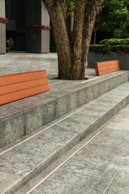 İnsanlar, içinde iki ahşap bank, bir ağaç ve taş basamakların bulunduğu bir park alanının önünden geçiyor. Sahne, arka planda binalar olan bir şehirde. Gündüz oldu..