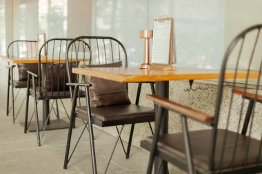 Açık bir kafe alanında metal sandalyeli birkaç ahşap masa kurulmuştur. Her masada küçük bir menü tutucu var. Sahne parlak ve temiz, müşteriler için rahat bir atmosfer gösteriyor..