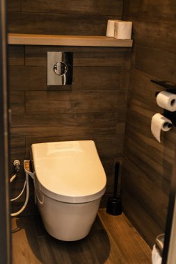 Küçük bir banyo ahşap bir rafın yanında modern bir tuvalet içerir. Rafta, tuvalet malzemeleri var. Bir rulo tuvalet kâğıdı yakındaki bir tutacakta ve tuvalet fırçası da tuvaletin yanında..