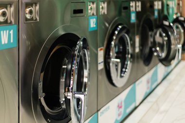 Makineler umumi bir çamaşırhanede duvara dizilmiş. Her birimin yuvarlak bir kapısı var. İnsanlar elbiselerini yükleyip yıkanmaya başlayabilir. Dükkanı müşteriler için bir sürü yer dolduruyor..