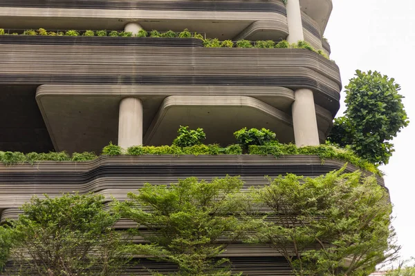 Sıralar halindeki bitkiler yüksek bir binanın balkonlarını süslüyor. Yapının çizgileri ve sütunları olan eşsiz bir tasarımı var. Binanın önündeki ağaçlar manzaraya yeşillik katıyor..