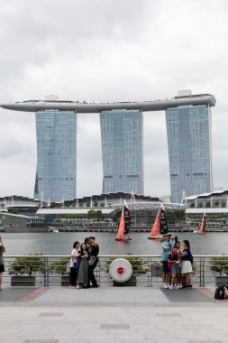 Singapur - 27 Ocak 2026: Marina Körfezi 'ndeki yürüyüş yolu boyunca turist grupları duruyor. Marina Bay Sands otel kuleleri arka planda yelkenliler suda ilerlerken. İnsanlar fotoğraf çeker.