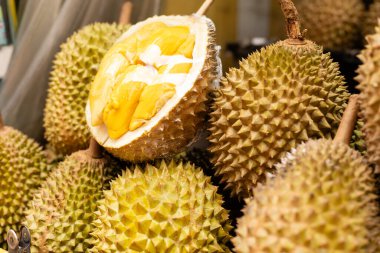 Durian meyveleri bir markette bir masanın üzerinde düzenlenir. Bir Durian kesilerek açılmış ve içindeki sarı et ortaya çıkmış. Ortam kalabalık görünüyor, tipik bir yerel piyasa sahnesi..