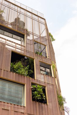 Modern bir bina, balkonlarında yeşilliklerle ayakta duruyor. Küçük bitkiler ve ağaçlar açık pencerelerden görülebilir. Gökyüzü parlak, şehir bölgesinde tipik bir günü gösteriyor..