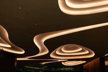 Modern bir binada kıvrımları akan ve yumuşak ışıkları olan bir tavan. Tasarım, geceleri mimariyi ön plana çıkaran eşsiz bir atmosfer yaratıyor..