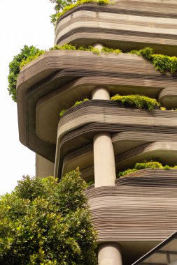 Yüksek bir binada çeşitli yeşil bitkilerle kaplı katmanlı balkonlar vardır. Yapı benzersiz kıvrımlar ve çizgiler gösteriyor. Güneş ışığı binanın üzerinde parlıyor, tasarımını vurguluyor..