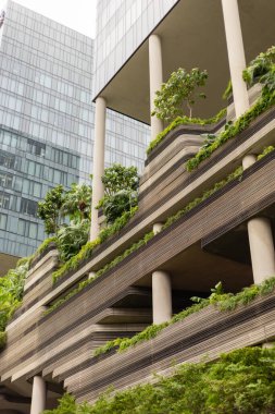 Yeşil bitki ve ağaçların yer aldığı birden fazla katlı bir bina yüksektir. Yapıda beton ve bitki örtüsü karışımı var. Günışığı modern tasarımı vurgular.