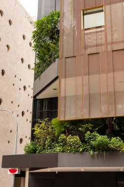 Çatısında ve girişinde yeşil bitkiler olan bir caddenin köşesinde bir bina duruyor. Yapısı modern ve metal elementlere sahip. Yakınındaki duvarlar desenli ve açık renkli..