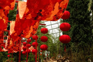 Kırmızı fenerler gün boyunca bir bahçe ortamında sallanırken sancaklar yukarıda asılı. Yeşil bitkiler görünür, ziyaretçiler için şenlikli bir atmosfer yaratıyor..