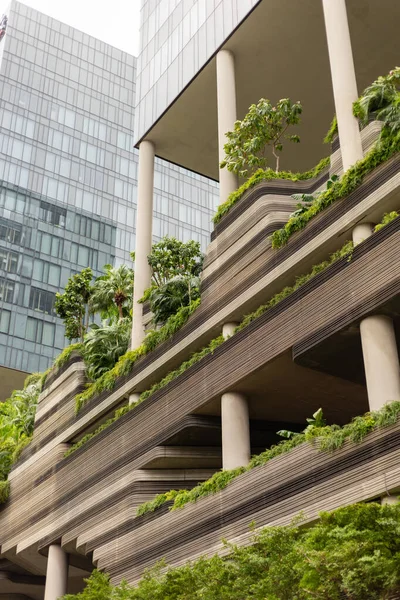 Yeşil bitki ve ağaçların yer aldığı birden fazla katlı bir bina yüksektir. Yapıda beton ve bitki örtüsü karışımı var. Günışığı modern tasarımı vurgular.