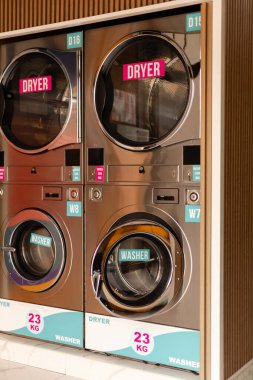 Çamaşır odasında, çamaşır ve kurutma makineleri içinde giysiler varken çalışır. İnsanlar çamaşırlarının bitmesini bekliyor. Makineler modern ve geniş boyutludur, açık etiketler gösterirler.