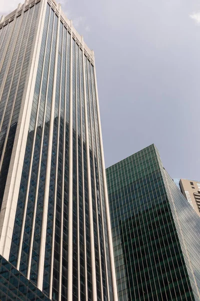 İki yüksek bina birbirine yakın duruyor. Birinin dikey çizgileri ve parlak bir yüzeyi var. Diğerinde yeşil cam bir cephe var. Gökyüzü mavi ve gündüz bulutları var..