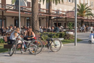 Bir sahil kasabasının işlek rıhtım manzarası. İki genç kız konuşuyor, bir bankta oturuyorlar, bisikletleri yanlarında duruyor, arka planda insanlarla dolu açık bir kafe manzarası var.