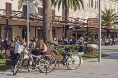 Bir sahil kasabasının işlek rıhtım manzarası. İki genç kız konuşuyor, bir bankta oturuyorlar, bisikletleri yanlarında duruyor, arka planda insanlarla dolu açık bir kafe manzarası var.