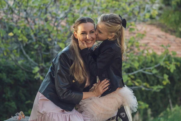 Mom and daughter take a walk in the park in autumn, both wearing pink dresses and black leather jackets, blondes with long hair. Little daughter hugs mom and whispers something in her ear