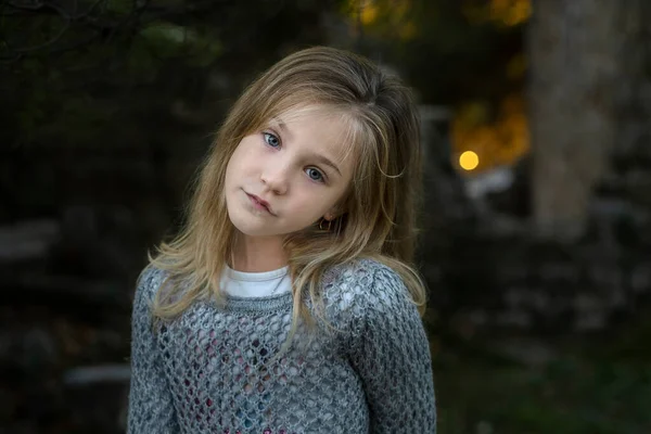 秋の公園を歩くグレーのニットセーターで長いブロンドの髪をした7歳の女の子の暗い背景に肖像画を閉じます — ストック写真