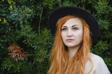 Siyah şapkalı ve gülümseyen kızıl saçlı güzel bir kızın portresini yakından çek. Arka planda şehir parkının yeşil çalıları var.