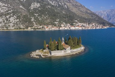 Hava görüntüsü, insansız hava aracı mavi denizin üzerinden küçük adaya uçuyor. Bir adada Hıristiyan kilisesi, konut binaları ve kozalaklı ağaçlar var. Arka planda dağlar ve kıyı şeridi görülebilir.