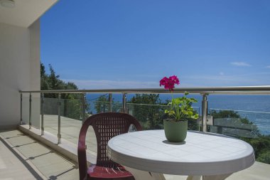 Deniz manzaralı özel villada geniş bir teras. Teras, yeşilliklerle kaplı deniz ve kıyı şeridi manzarası sunuyor. Terasta sandalyeli plastik bir masa var. Masanın üzerinde de dekorasyon için kırmızı bir çiçek var.