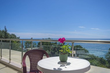 Deniz manzaralı özel villada geniş bir teras. Teras, yeşilliklerle kaplı deniz ve kıyı şeridi manzarası sunuyor. Terasta sandalyeli plastik bir masa var. Masanın üzerinde de dekorasyon için kırmızı bir çiçek var.