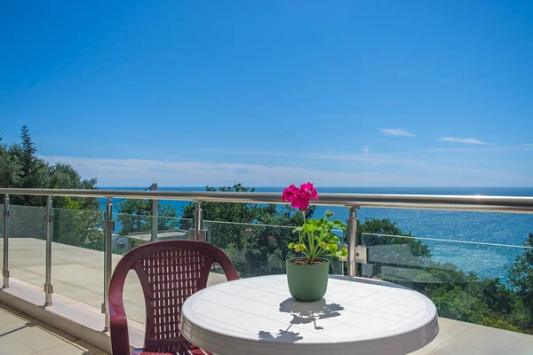 Deniz manzaralı özel villada geniş bir teras. Teras, yeşilliklerle kaplı deniz ve kıyı şeridi manzarası sunuyor. Terasta sandalyeli plastik bir masa var. Masanın üzerinde de dekorasyon için kırmızı bir çiçek var.