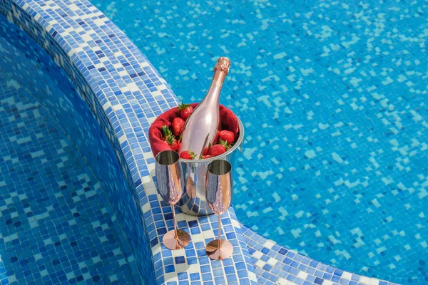 Swimming pool view. Close-up of a bucket with champagne, strawberries and two empty glasses on the side of the pool