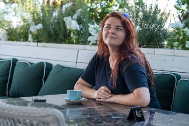 Açık hava kafedeki bir masada oturan uzun saçlı, gülümseyen kızıl saçlı genç bir kadının portresi. Akıllı telefon onun önünde, masanın üzerinde yatıyor. 