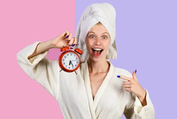Smiling morning woman with alarm clock. Happy girl in bathrobe with alarm watch. Woman pointing on alarm-clock. Time concept. — Stock Photo, Image
