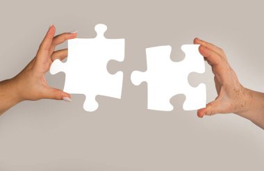 Gri arkaplandaki yapboz parçasını birleştirmeye çalışan iki el. Takım çalışması konsepti. Yapbozu birbirine bağlayan el. Bulmaca parçaları yaşlı ve genç elleri birbirine bağlıyor. Nesiller arasında bulmaca çözümü