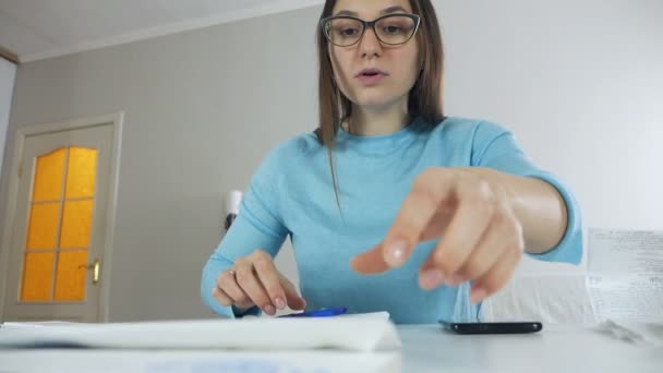 Een meisje zit aan een tafel in een appartement, onderzoekt nutsrekeningen en somt de resultaten op, geld beheert financiën, het familiebudget — Stockvideo