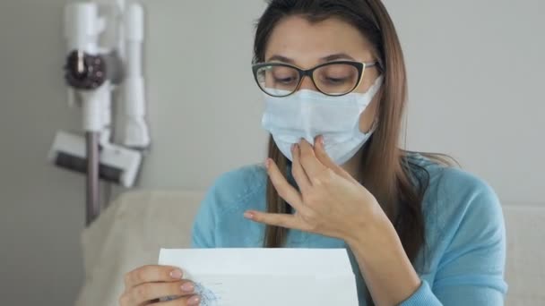 Une jeune femme s'assied à une table, avec un masque sur le visage, pendant la cohabitation, tenant et léchant une enveloppe blanche pour envoyer — Video