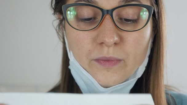 Eine junge Frau sitzt an einem Tisch, mit einer Maske auf dem Gesicht, beim Kochen, hält und leckt einen weißen Umschlag für den Versand — Stockvideo