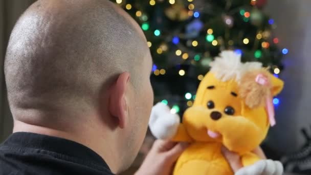 Um homem segura um ursinho, perto de uma árvore de Natal. conceito de pessoas esquisitas. brincar com um brinquedo. brinquedo calmante para o autismo — Vídeo de Stock