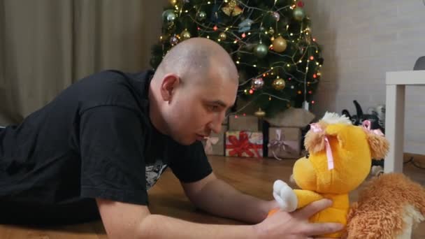 Een man houdt een teddybeer vast bij een kerstboom. Vreemde mensen concept. spelen met een speeltje. verzachtend speelgoed voor autisme — Stockvideo