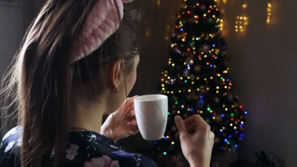 Romantikus lány egy csésze kávéval vagy teával. Gyönyörű lány élvezi a kávét. Ünnepi belső tér fényekkel, karácsonyfa Téli este, hangulatos