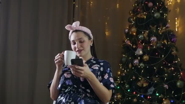 Romantyczna dziewczyna z filiżanką kawy lub herbaty. Piękna dziewczyna lubi kawę i korzysta z telefonu. Uroczyste wnętrze ze światłami, Choinka Zimowy wieczór, przytulne — Wideo stockowe