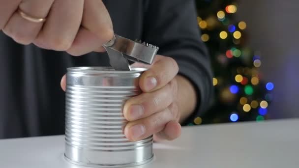 Primer plano de fuertes manos masculinas con abrelatas haciendo una incisión en la tapa de una lata de metal frente al árbol de navidad — Vídeos de Stock