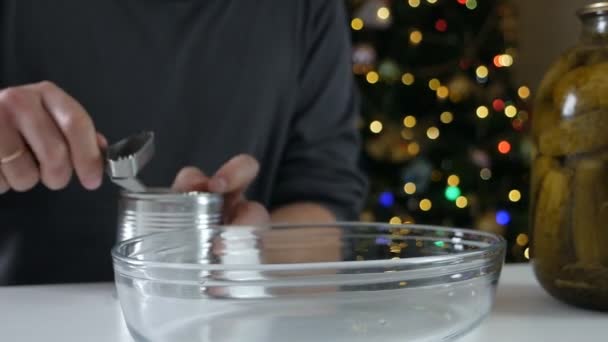 Hombre fuerte utilizando las manos abrelatas cortar la tapa de una lata de metal en una mesa con encurtidos en frente de un árbol de navidad.guisantes verdes para ensalada tradicional — Vídeos de Stock